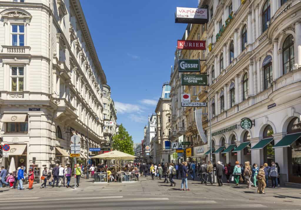 Bustling daytime scene on a street in Vienna with pedestrians, outdoor cafes, and classic Viennese architecture lining the road