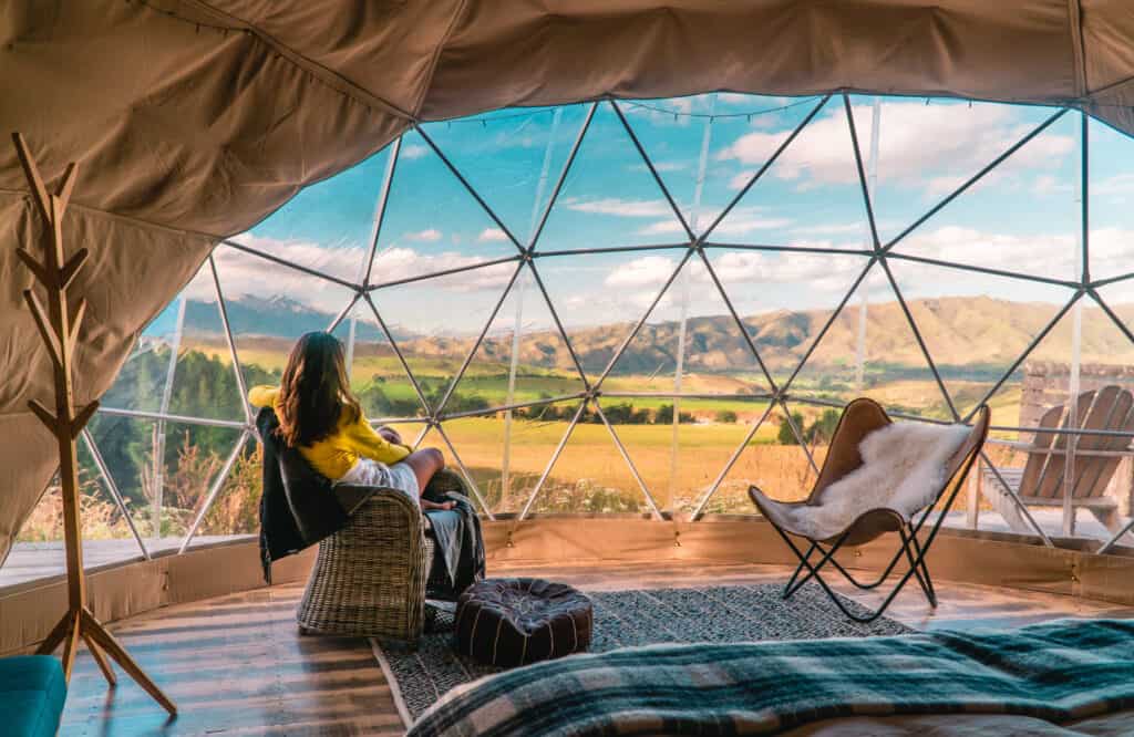 A person sitting inside a geodesic dome tent with a view of rolling hills and a clear sky, exemplifying serene glamping