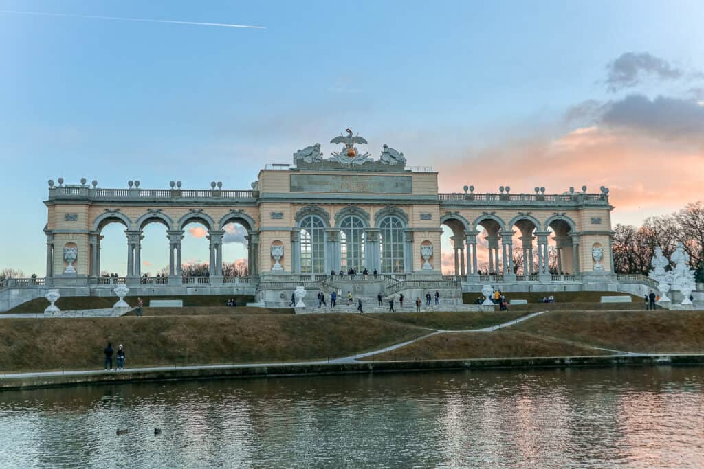 "Sunset at the Gloriette in Vienna, with the structure's arches silhouetted against a pink-tinged sky reflecting off a calm pond