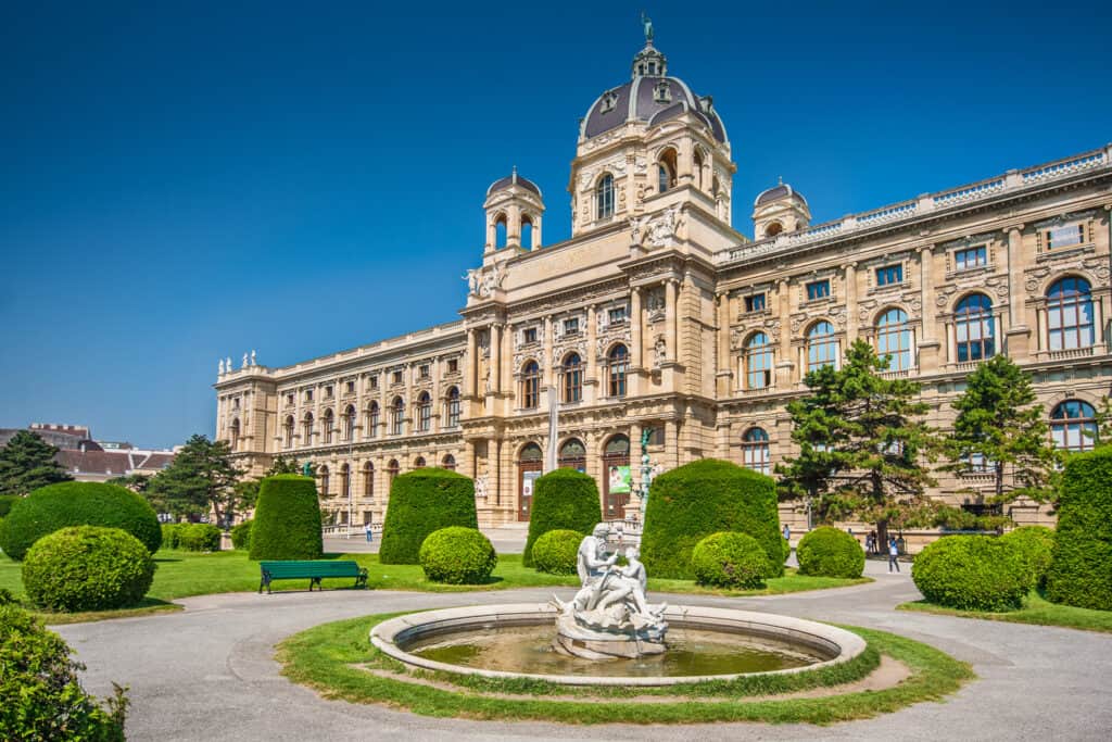 Vienna's Natural History Museum fronted by manicured hedges and a classical statue, on a bright, sunny day with a clear blue sky