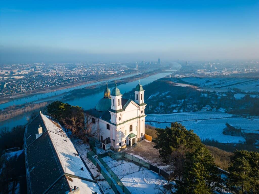 The historic Leopoldsberg church overlooking a frost-covered Vienna with the Danube River splitting the cityscape under a pastel morning sky