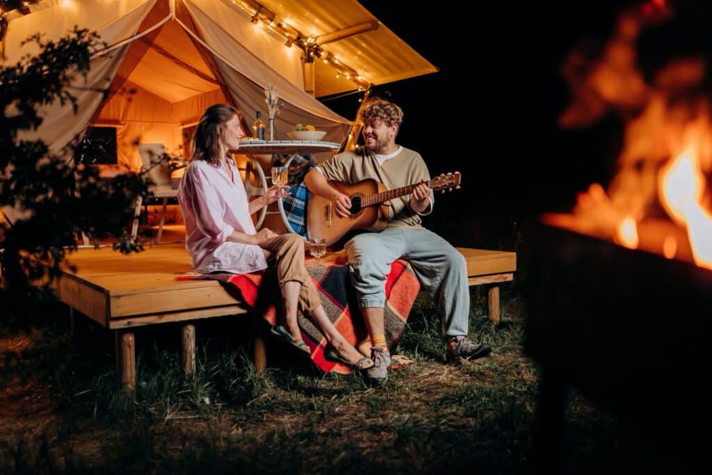 A couple relaxing near a glamping tent at night, with a man playing guitar and a woman enjoying a drink by the fireside