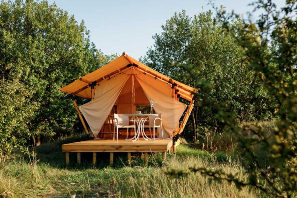 A glamping tent on a wooden platform surrounded by lush greenery, featuring a welcoming set of chairs and a table