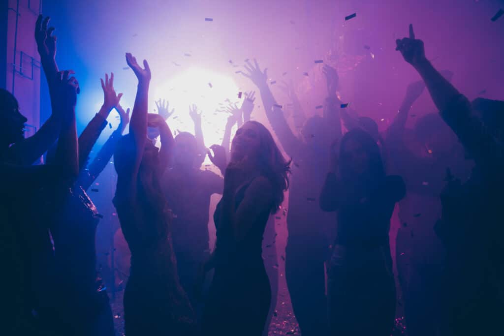 A silhouette of a joyous crowd with hands raised, celebrating at a party under purple lights and flying confetti