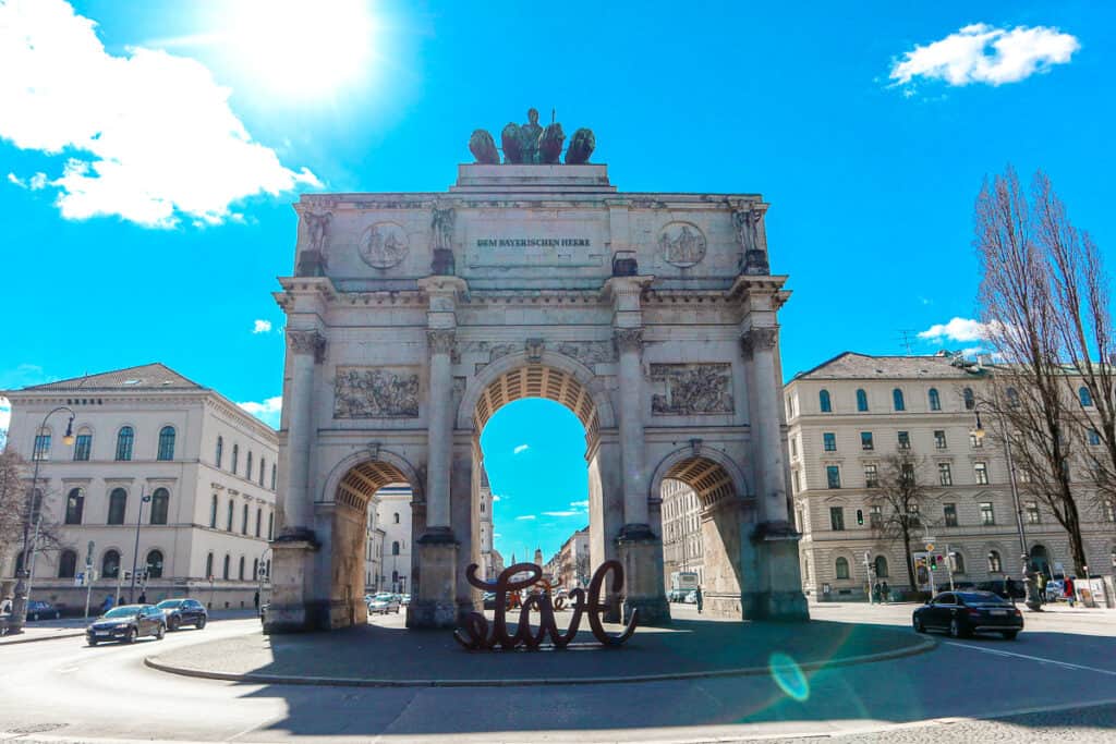 The iconic Siegestor (Victory Gate) in Munich with a modern sculpture spelling 'love' in the foreground under a bright blue sky