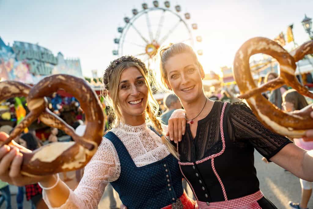 Two women in traditional Bavarian dresses holding pretzels in front of a Ferris wheel at Oktoberfest in Munich