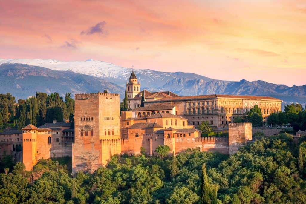 The Alhambra, a majestic fortress complex, is bathed in the warm glow of sunset, with the snow-capped Sierra Nevada mountains in the distance.