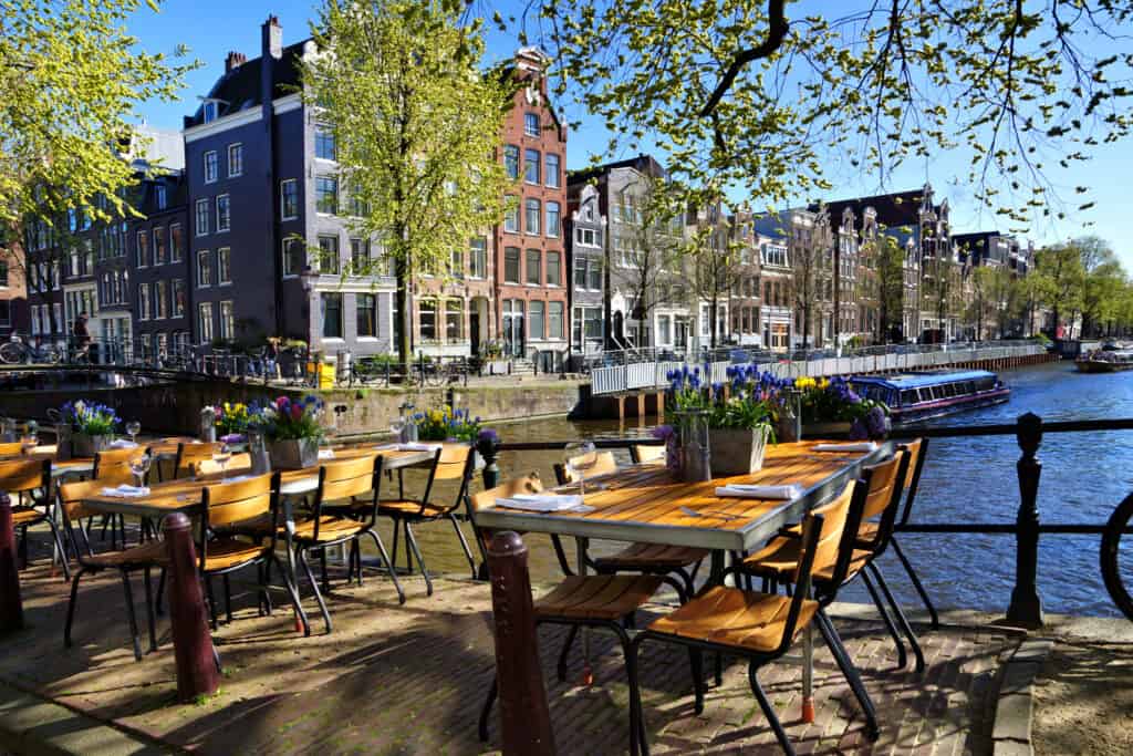 Outdoor riverside caf in Amsterdam with wooden tables set up for dining, overlooking the beautiful canal and passing boats