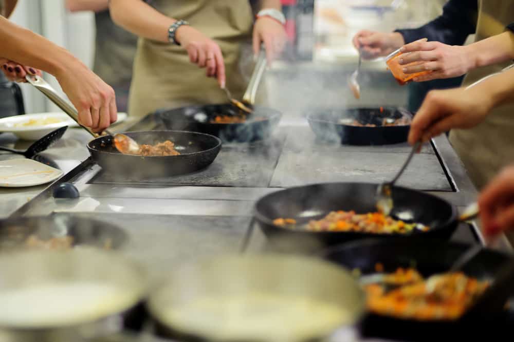 An engaging cooking class in action with multiple chefs sauting dishes in pans over a stove, creating a dynamic culinary atmosphere.
